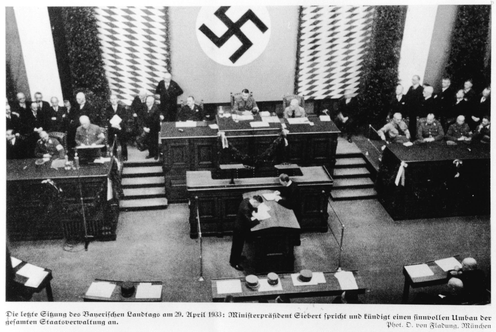Abgeordnete tagen 1933 im Bayerischen Landtag. Das Hakenkreuz der Nationalsozialisten hängt im Hintergrund großflächig an der Wand.